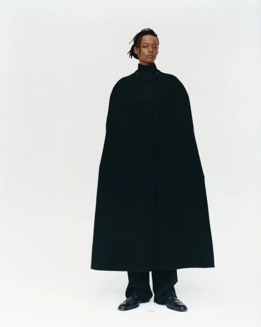 Young adult with locks stands in a photo studio wearing a black turtleneck, cape, pants and shoes against a white backdrop.