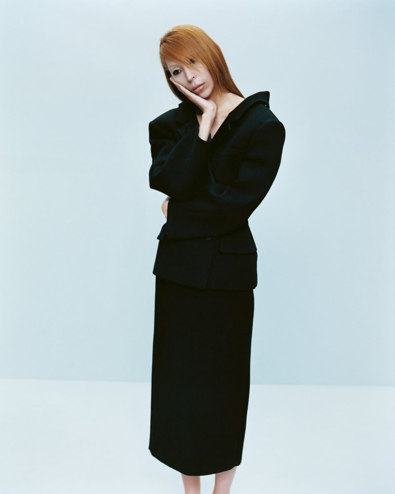 Full-length portrait of an adult woman with long copper hair, wearing a black suit, posing against a light blue studio background.
