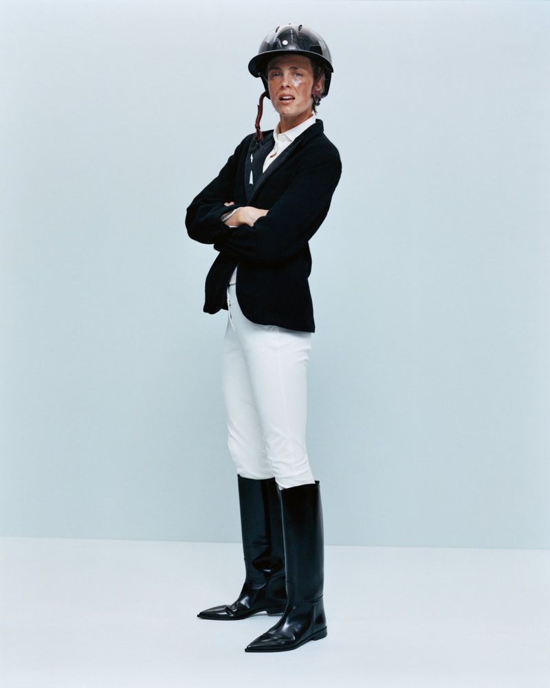 A person wearing equestrian attire, including a helmet, jacket, white pants, and black boots, poses with arms crossed against a light blue backdrop.