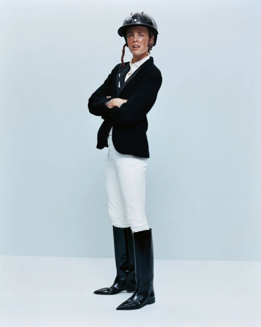 A person wearing equestrian attire, including a helmet, jacket, white pants, and black boots, poses with arms crossed against a light blue backdrop.