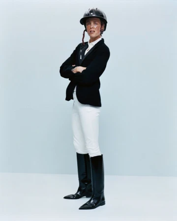 A person wearing equestrian attire, including a helmet, jacket, white pants, and black boots, poses with arms crossed against a light blue backdrop.