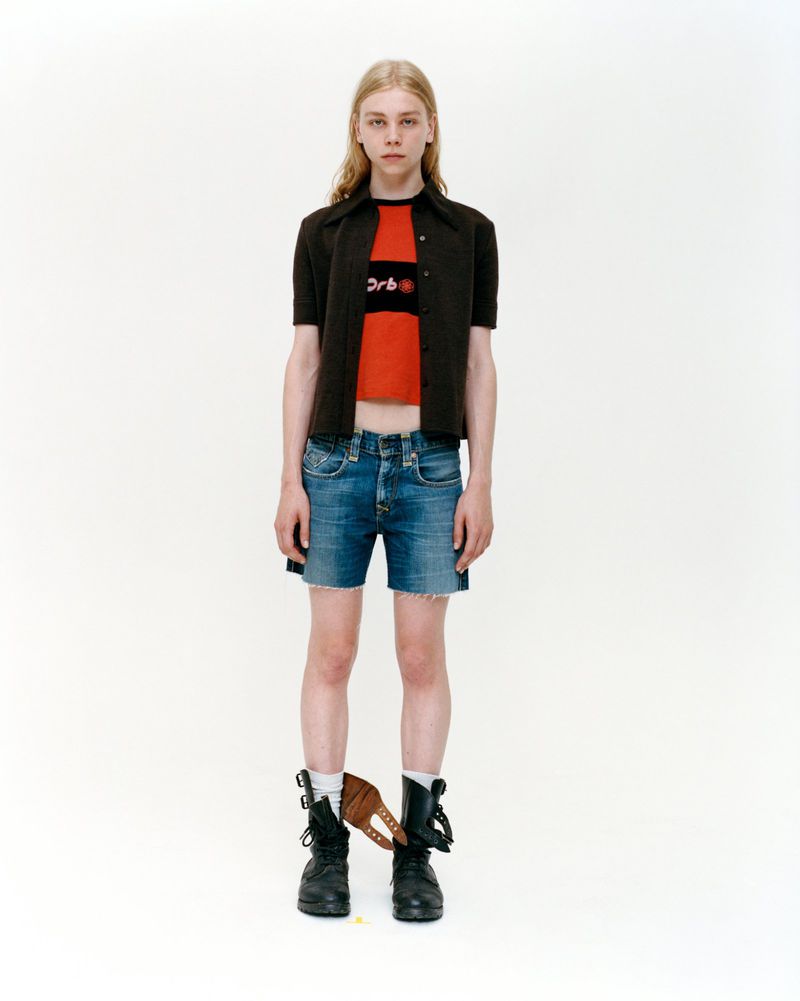 Full-length portrait of a young adult with long blonde hair, wearing a red top, brown cardigan, blue shorts, and black boots in studio.