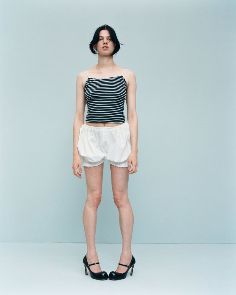 Standing woman in a striped top, white shorts, and black heels against a light blue backdrop in a full shot.