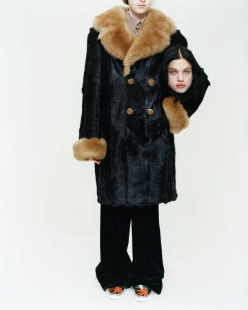 Fashion model in a black fur coat with tan shearling collar holding a head under one arm against a white studio background.