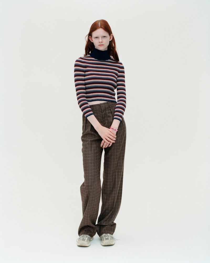 Full shot of a young red-haired woman in a horizontally striped long-sleeve shirt and plaid trousers, standing against a white backdrop.