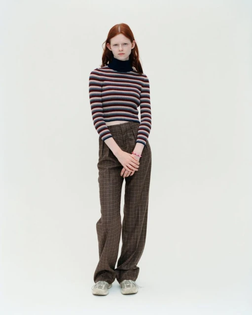 Full shot of a young red-haired woman in a horizontally striped long-sleeve shirt and plaid trousers, standing against a white backdrop.