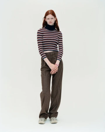 Full shot of a young red-haired woman in a horizontally striped long-sleeve shirt and plaid trousers, standing against a white backdrop.