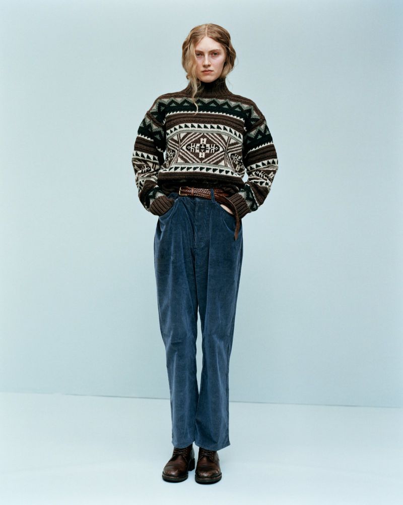 Model wearing a patterned turtleneck sweater and blue corduroy trousers against a neutral backdrop.