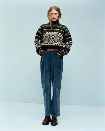 Model wearing a patterned turtleneck sweater and blue corduroy trousers against a neutral backdrop.