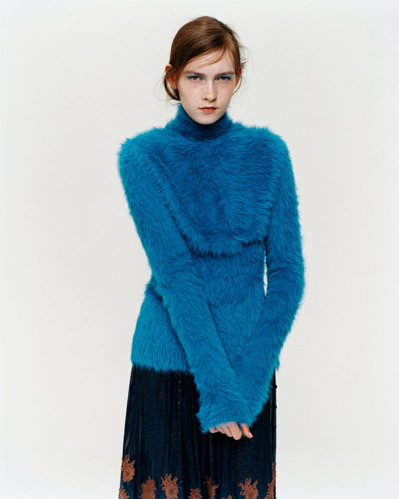 Woman in a textured blue sweater and patterned skirt poses in a studio. The subject is centered, with a bright and minimalist background.