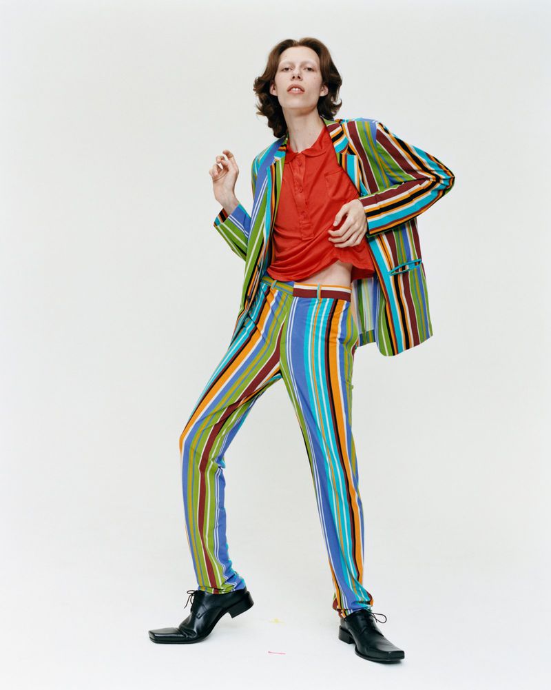 Person in colorful striped suit and black shoes poses on a white background, looking slightly upward, fashion-forward style, studio lighting.