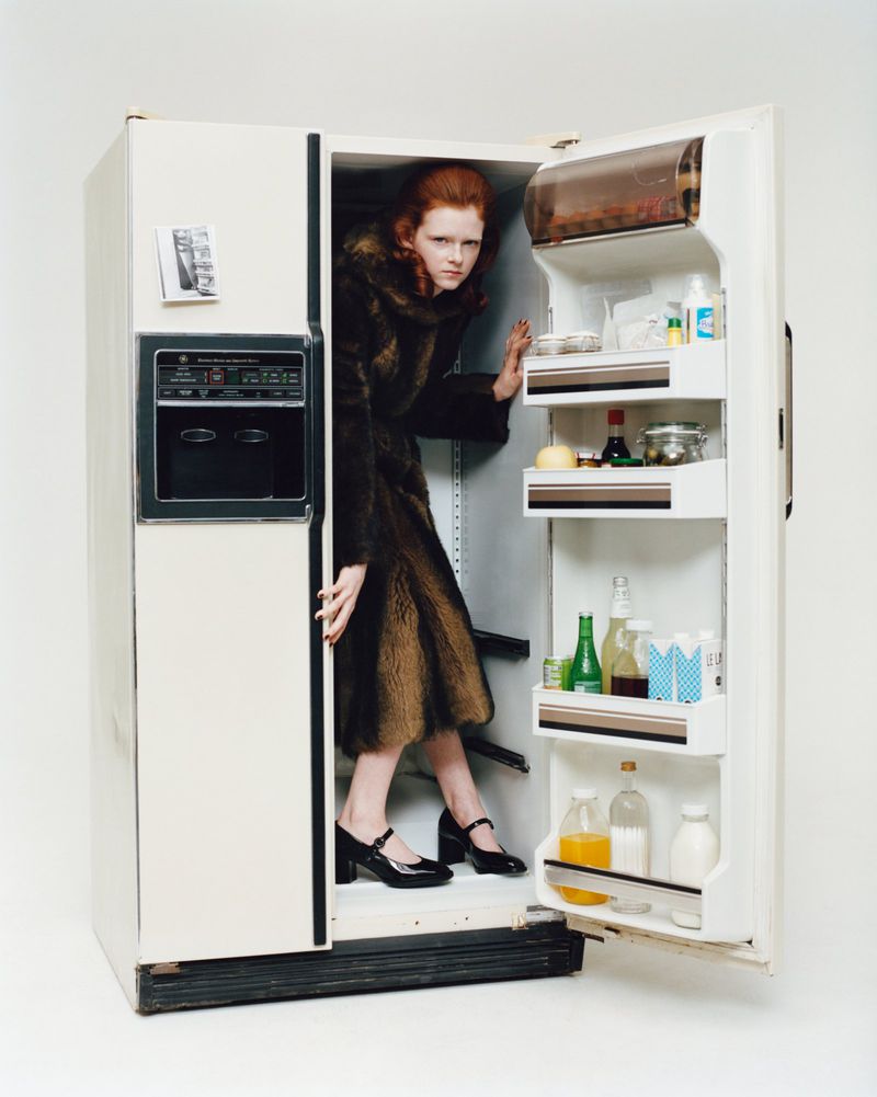 A red-haired woman in a fur coat stands inside an open vintage refrigerator stocked with various bottles and containers, with a white background.