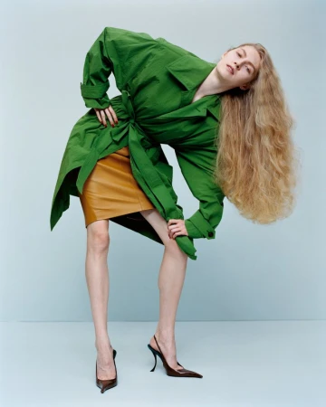Fashion portrait of a woman in a green jacket and brown skirt bending backwards against a light blue background in a studio.
