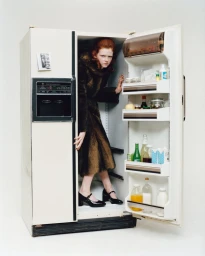 Red-haired model standing inside a large open white refrigerator wearing a dark faux fur coat and black Mary Jane shoes.