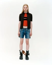 Full-length portrait of a model in a dark button-up shirt, red top, and denim shorts against a white background.