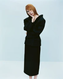 Model with reddish-brown hair wearing a structured black blazer and matching skirt against a white studio background.
