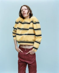 Model wearing a yellow and black striped cropped sweatshirt and burgundy trousers against a light blue backdrop.