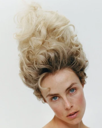 Close-up portrait of a woman with an exaggerated, voluminous high blonde bouffant hairstyle against a plain white background.
