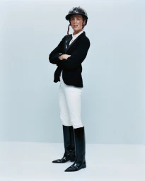 Equestrian model wearing a black riding jacket, white trousers, and tall boots with a helmet against a light grey background