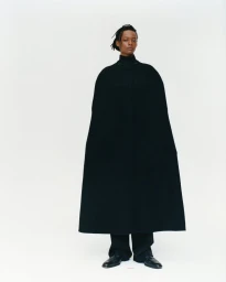 Model standing in a long structured black cape and dark trousers against a clean white studio backdrop.