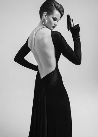 Model in a black backless gown holding a handgun against a plain grey seamless background for 5ELEVEN Magazine