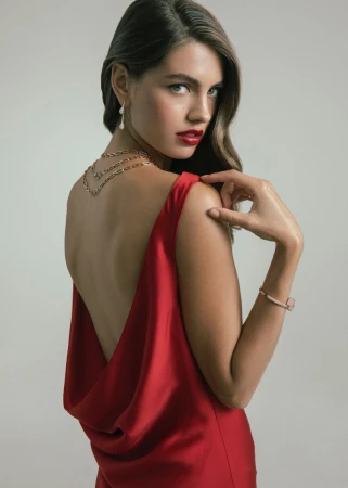 Woman looking over her shoulder wearing a backless red cowl drape dress, gold chain necklace, and bold red lipstick against a studio backdrop.