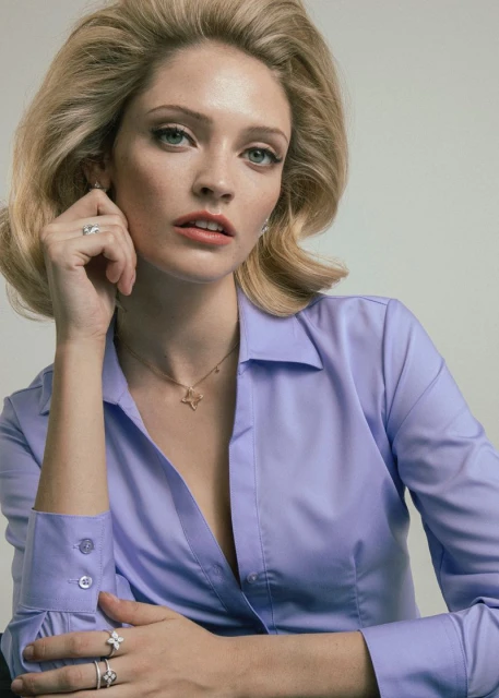 Close-up portrait of a blonde woman wearing a periwinkle button-down shirt and jewelry indoors in a photo studio with a neutral background.