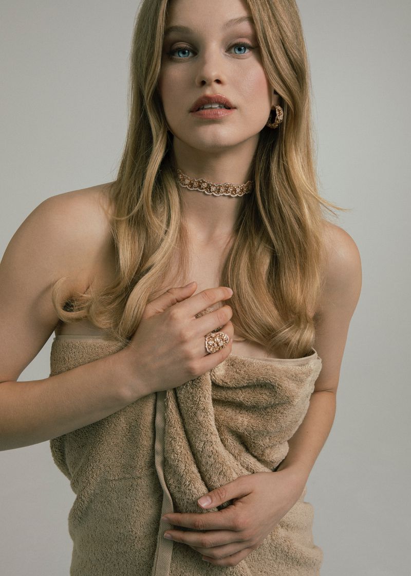 Young woman wrapped in a towel wearing jewelry poses in a studio with a neutral background, showcasing fashion and beauty elements.