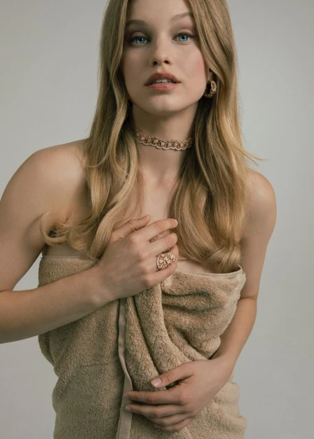 Young woman wrapped in a towel wearing jewelry poses in a studio with a neutral background, showcasing fashion and beauty elements.
