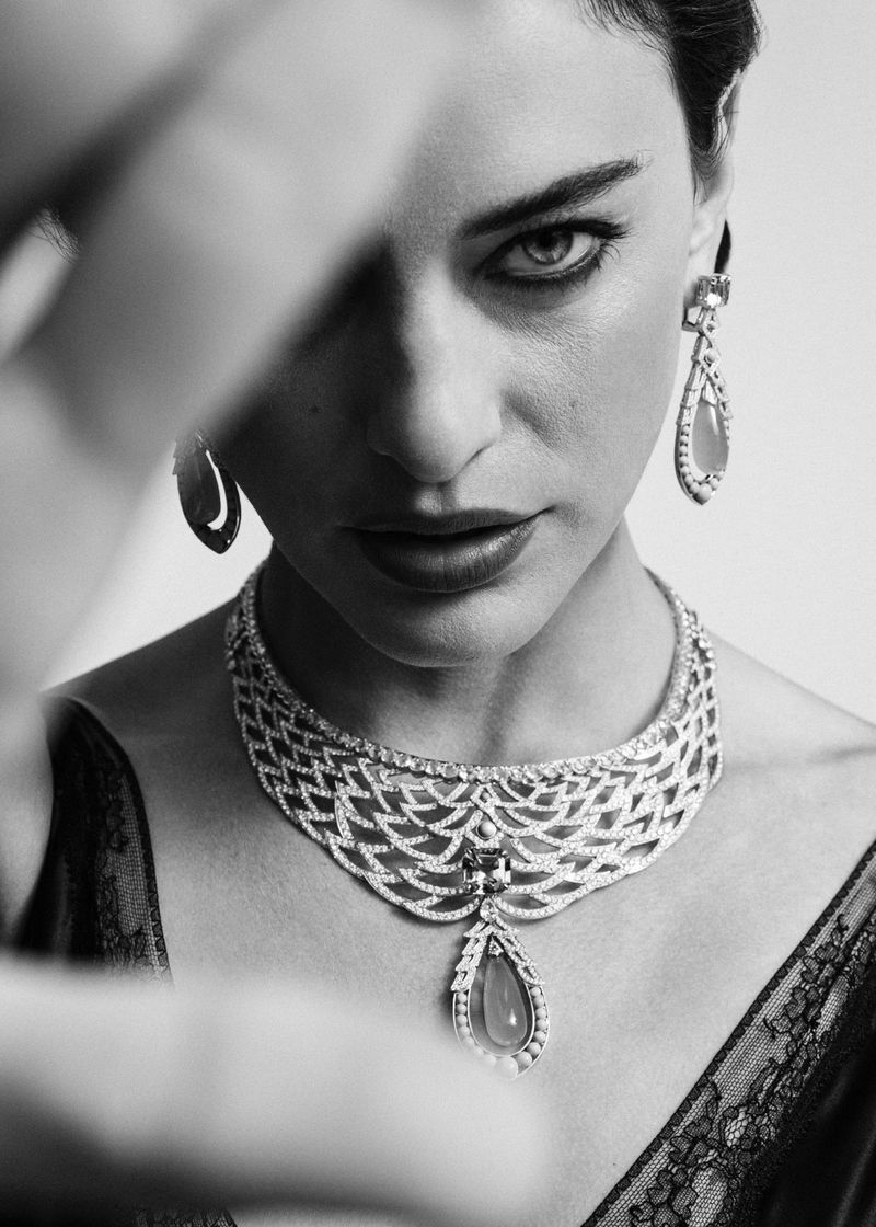 Black and white portrait of a woman wearing a statement necklace and earrings, looking at the camera with a hand partially obscuring her face.