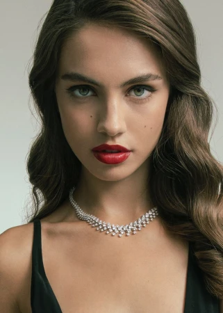 Close-up of a beautiful adult woman with red lipstick and wavy brown hair wearing a diamond necklace against a neutral background.