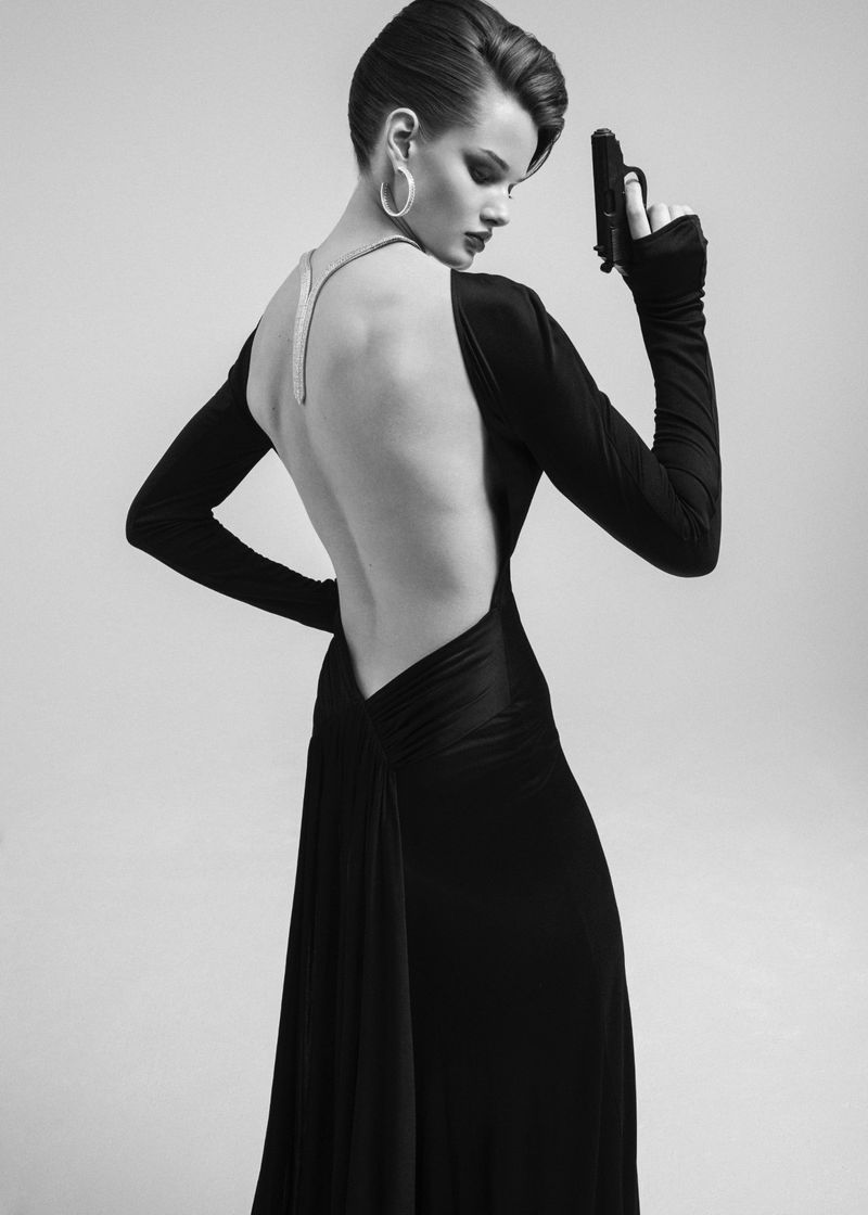 Woman in elegant black dress holding a handgun, posing in a photo studio with a simple grey backdrop and soft lighting.