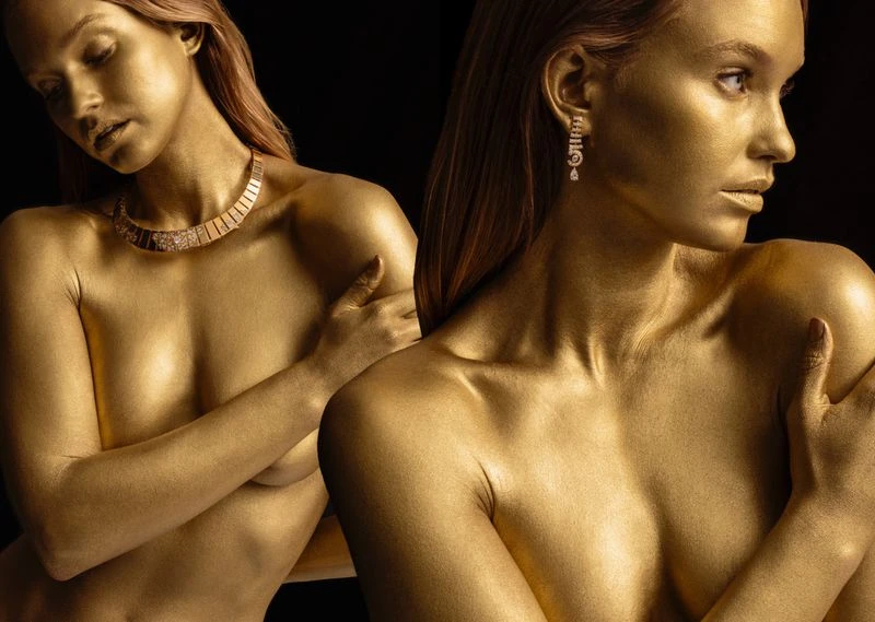 Two women with golden skin and makeup pose in front of a black background, showcasing jewelry and a fine art photography style.