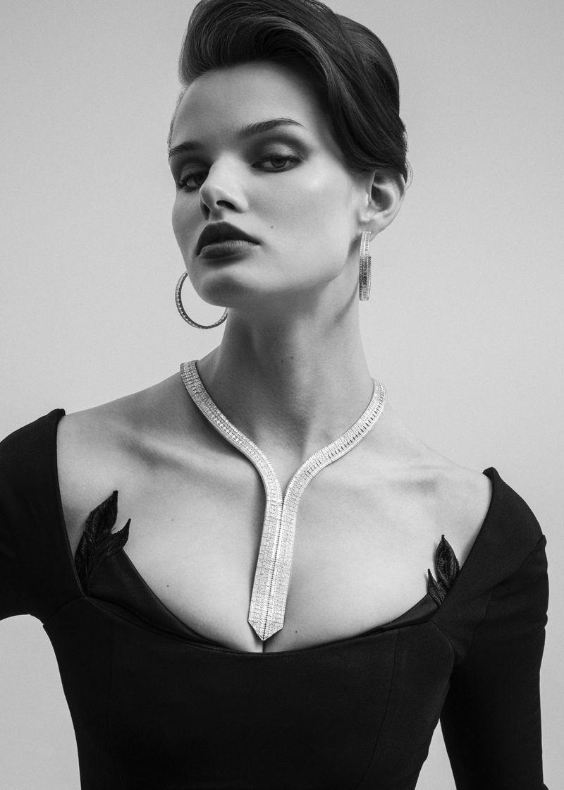 Black and white portrait of an adult female model wearing diamond jewelry including a necklace and hoop earrings looking at the camera.