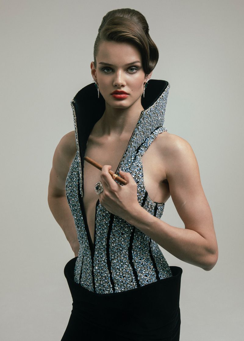 Glamorous woman in shimmering top holding a cigar in a studio setting. Sophisticated hair and makeup complete the high-fashion look.