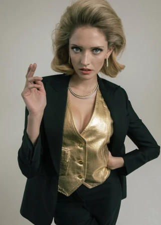 A blonde woman poses in a studio wearing a black suit jacket and trousers, with a gold vest. She has green eyes and holds up a finger.