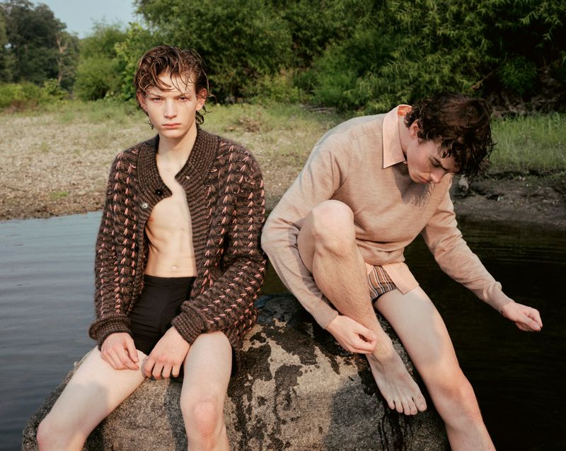 Two fair-skinned men sitting on a rock by a river, one wearing a cardigan, the other a collared shirt, in a natural outdoor setting.