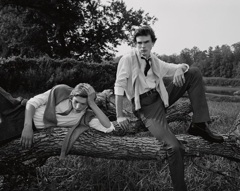 Two young men lounge on a fallen log in a grassy field in a black and white outdoor shot, both wearing collared shirts.