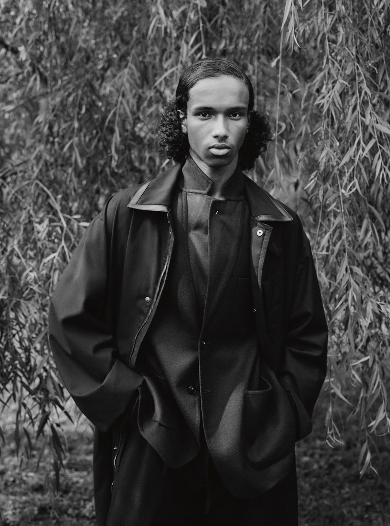 Portrait of a young adult male model in a dark jacket and matching pants, standing outdoors against a weeping willow tree.