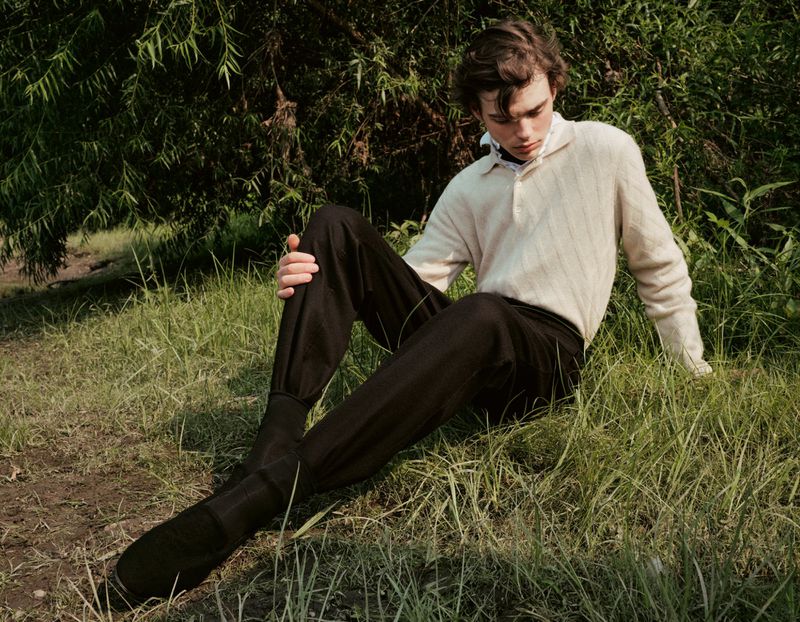 Young adult male sits in a field of grass wearing black pants and cream shirt. Natural light illuminates this outdoor setting.