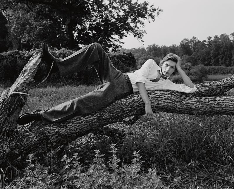 Monochrome portrait of a young adult male model reclining on a fallen tree in a grassy field wearing a white shirt and dark pants.