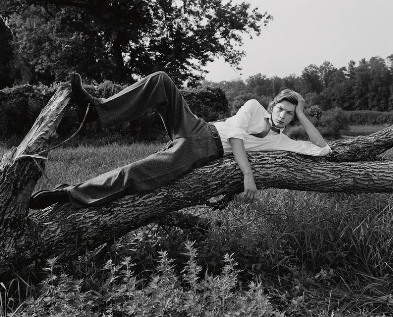 Monochrome portrait of a young adult male model reclining on a fallen tree in a grassy field wearing a white shirt and dark pants.
