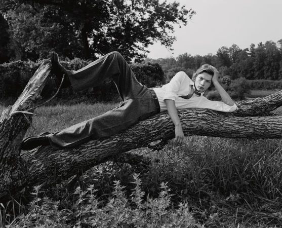 Monochrome portrait of a young adult male model reclining on a fallen tree in a grassy field wearing a white shirt and dark pants.