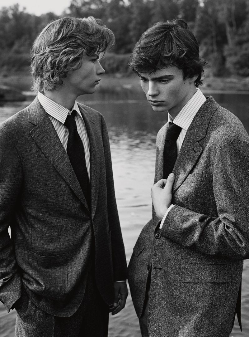Two men in tailored wool suits and collared shirts with ties standing outdoors beside a body of water in black and white.