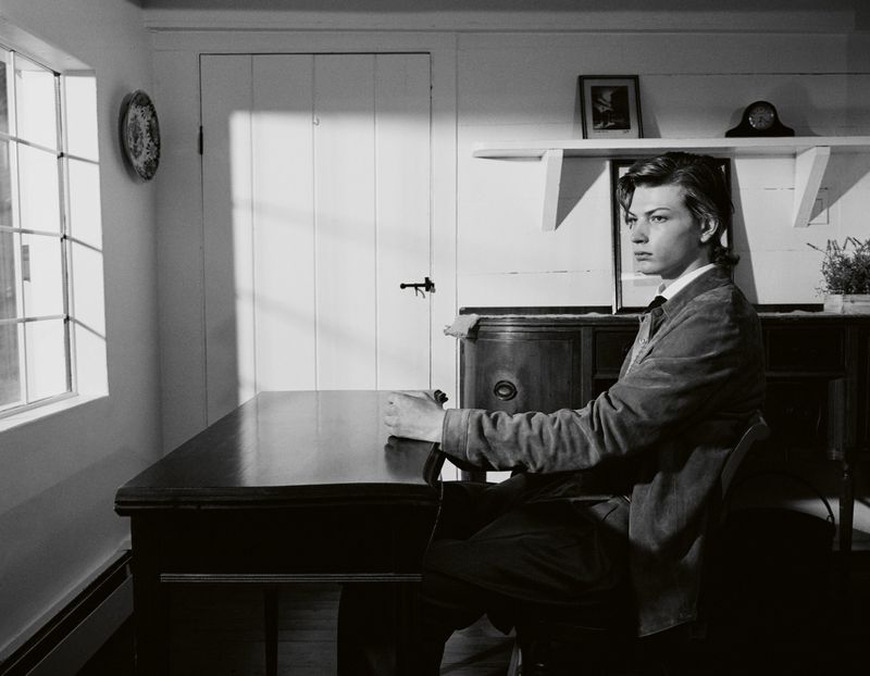 Black and white portrait of a pensive adult male in a suit jacket, sitting at a table in a domestic interior setting with a window.