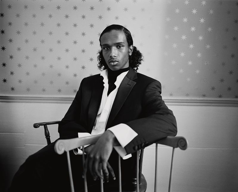 Sabir Abdikadir in a tailored black suit jacket and wide-cuff shirt seated on a wooden chair against star-patterned wallpaper.