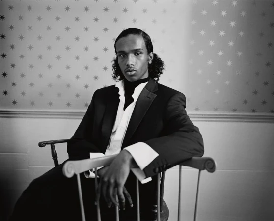 Sabir Abdikadir in a tailored black suit jacket and wide-cuff shirt seated on a wooden chair against star-patterned wallpaper.