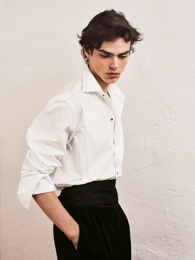 Portrait of a young man in a white shirt and black pants, looking down with a serious expression against a plain wall indoors.