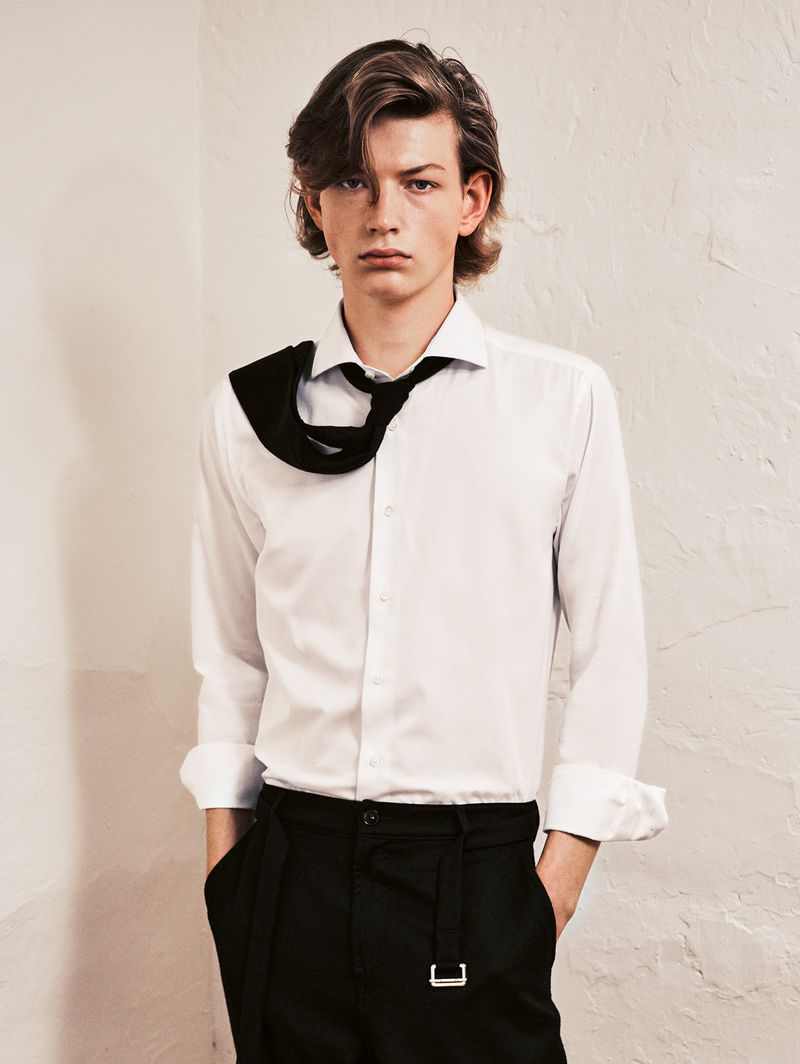 Studio shot of an adult male model with medium-length brown hair, wearing a white shirt and black pants in front of a textured off-white wall.