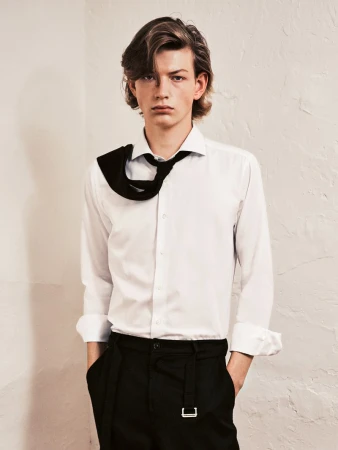 Studio shot of an adult male model with medium-length brown hair, wearing a white shirt and black pants in front of a textured off-white wall.